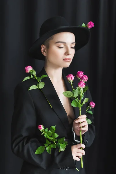 Young woman in black brim hat and blazer posing with closed eyes and pink roses on dark background — Stock Photo