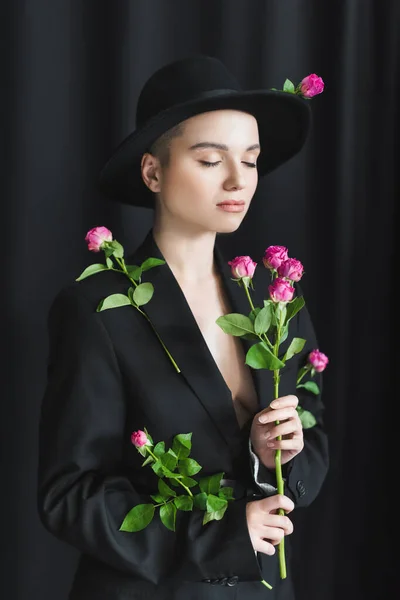 Mujer joven en sombrero de ala negra y chaqueta posando con los ojos cerrados y rosas rosadas sobre fondo oscuro - foto de stock