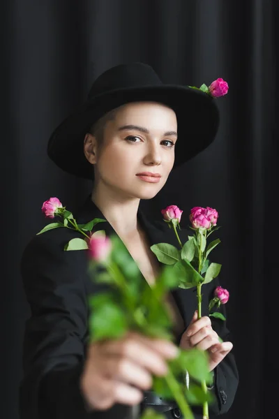 Jolie femme au chapeau bord et blazer noir tenant des roses roses sur fond sombre — Photo de stock