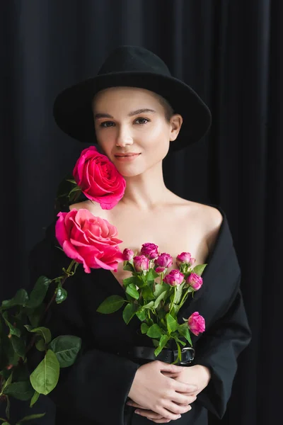 Mujer joven en chaqueta negra y sombrero de ala mirando a la cámara cerca de rosas frescas sobre fondo oscuro - foto de stock