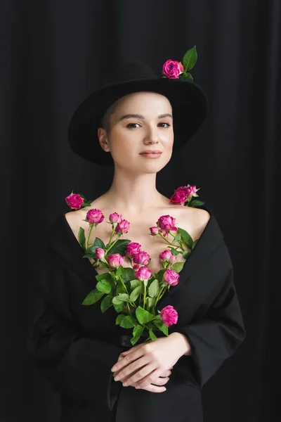 Charmante Frau in schwarzem Blazer und Kremphut posiert mit frischen rosa Rosen auf dunklem Hintergrund — Stockfoto