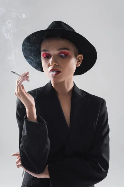 Frau mit schwarzem Fedora-Hut und Blazer raucht isoliert auf grau — Stockfoto