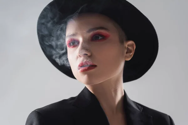 Retrato de mujer con maquillaje rojo carmín exhalando humo aislado en gris - foto de stock