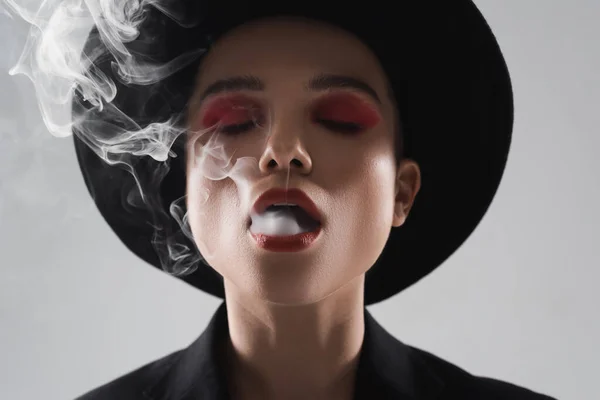 Front view of stylish woman with closed eyes exhaling smoke isolated on grey — Stock Photo