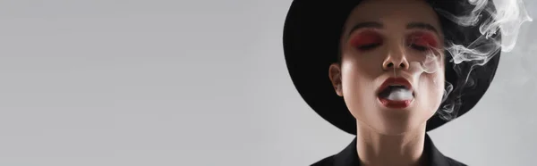 Mujer en sombrero de ala negra, con maquillaje brillante, exhalando humo aislado en gris, bandera - foto de stock