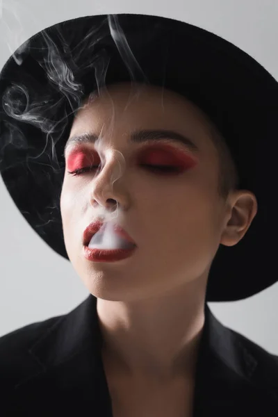 Mujer con los ojos cerrados y maquillaje rojo carmín exhalando humo aislado en gris - foto de stock