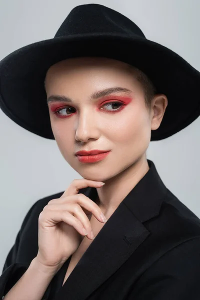 Jeune femme avec maquillage rouge carmin, chapeau bord noir, menton touchant isolé sur gris — Photo de stock