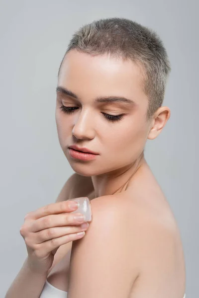Красивая женщина с чистой кожей нанося кубик льда на обнаженное плечо изолированы на серый — стоковое фото
