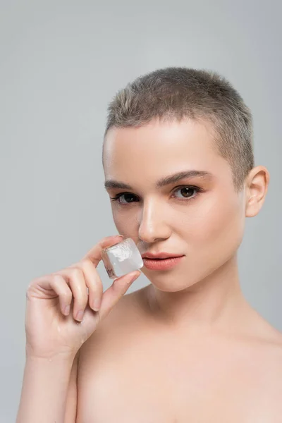 Jovem mulher com cabelo curto olhando para a câmera enquanto segurando cubo de gelo isolado em cinza — Fotografia de Stock