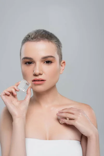 Mujer joven con pelo corto sosteniendo cubitos de hielo y mirando a la cámara aislada en gris - foto de stock