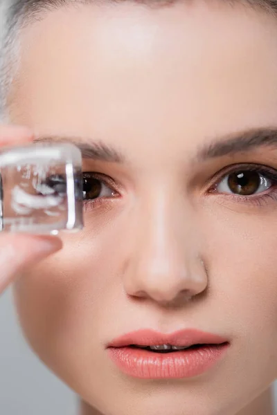 Vista de cerca de la cara femenina con la piel perfecta cerca del cubo de hielo aislado en gris - foto de stock