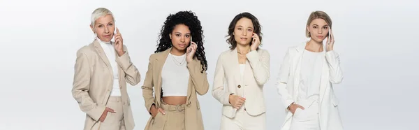 Multiethnic businesswomen in formal wear talking on cellphones isolated on grey, banner — Stock Photo