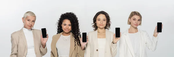 Interracial businesswomen in suits holding smartphones isolated on grey, banner — Stock Photo