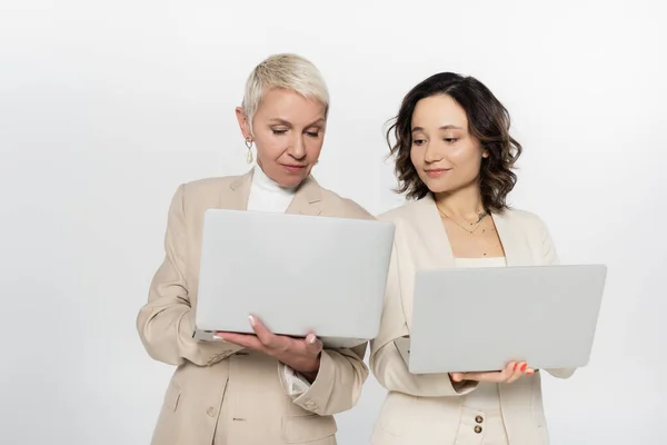 Senior businesswoman holding laptop near colleague isolated on grey — Stock Photo