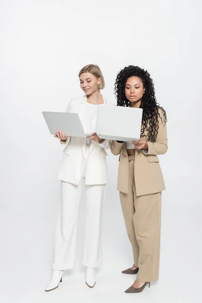 Full length of stylish interracial businesswomen using laptops on grey background — Stock Photo