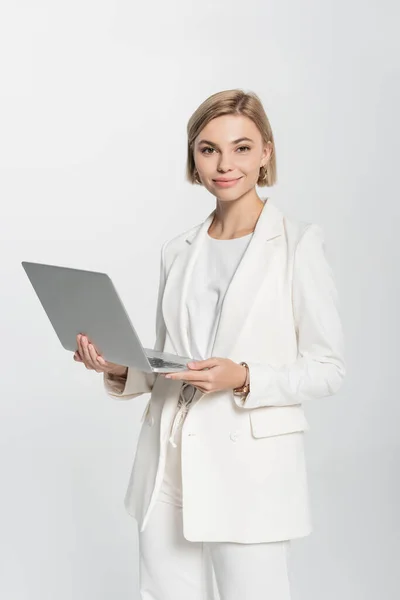 Donna d'affari sorridente in tuta che tiene il computer portatile e guarda la fotocamera isolata sul grigio — Foto stock