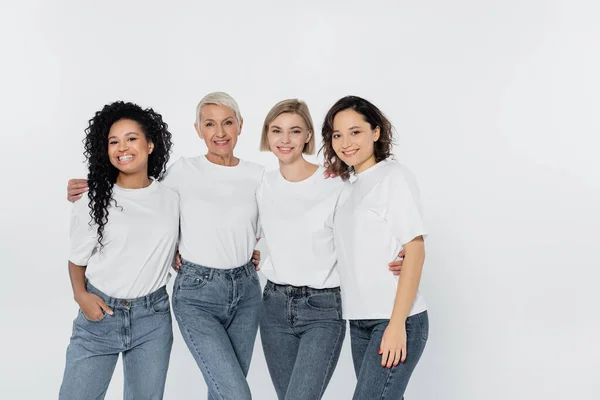 Glückliche multiethnische Frauen in weißen T-Shirts umarmen sich vereinzelt auf grau — Stockfoto