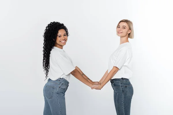 Mujeres jóvenes interraciales cogidas de la mano aisladas en gris - foto de stock