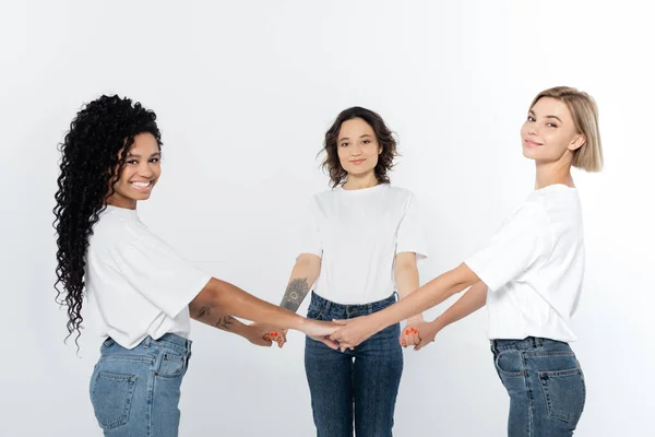 Positive multiethnische Frauen in weißen T-Shirts, die Hände isoliert auf grauem Feminismus-Konzept halten — Stockfoto