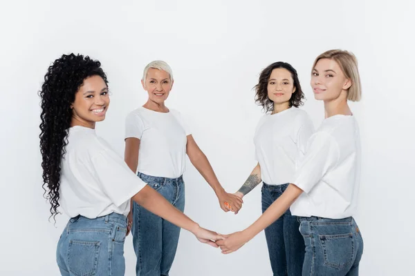 Multiethnic women smiling at camera and holding hands isolated on grey — Stock Photo