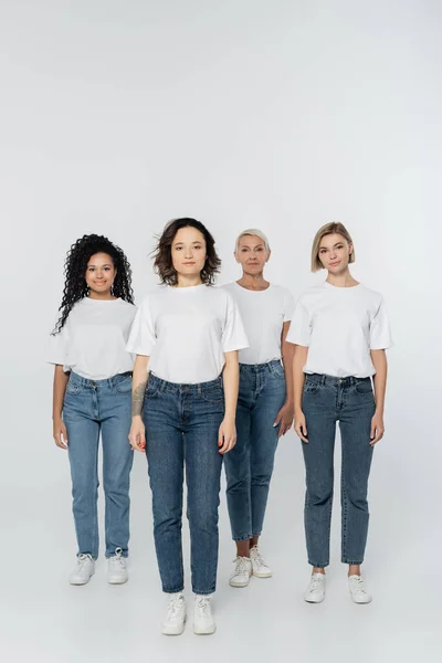 Full length of smiling multiethnic women in white t-shirts standing on grey background — Stock Photo