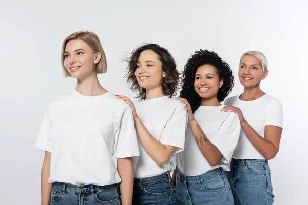 Mujeres sonrientes multiétnicas con camisetas blancas mirando hacia otro lado aisladas en gris - foto de stock