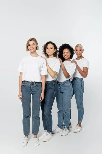 Longitud completa de mujeres multiétnicas en jeans y camisetas blancas mirando a la cámara sobre fondo gris - foto de stock