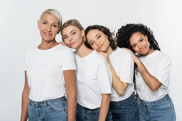 Mujeres jóvenes multiétnicas en camisetas blancas abrazándose cerca de una amiga sonriente aislada en un concepto de feminismo gris - foto de stock