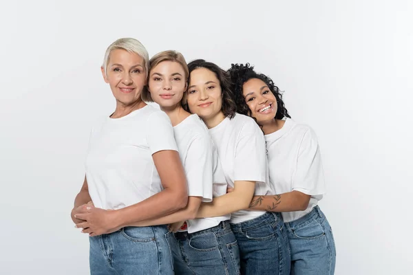 Alegre interracial mulheres no branco t-shirts abraçando cada outros isolado no cinza — Fotografia de Stock