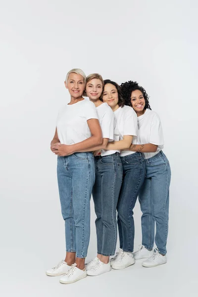 Comprimento total de mulheres multiétnicas sorridentes abraçando em fundo cinza, conceito de feminismo — Fotografia de Stock