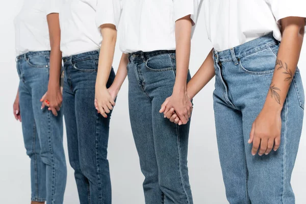 Vista recortada de mujeres interracial en camisetas y vaqueros tomados de la mano aislados en gris - foto de stock