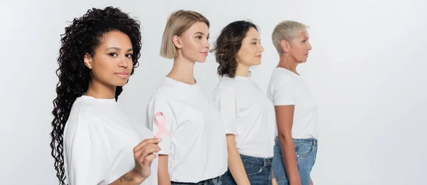 Young african american woman holding pink ribbon of breast cancer awareness near friends isolated on grey, banner — Stock Photo