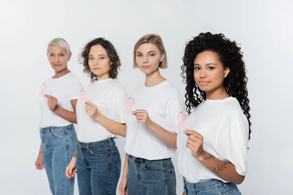 Mujeres interraciales sosteniendo cintas rosadas de conciencia sobre el cáncer de mama y mirando a la cámara aislada en gris - foto de stock