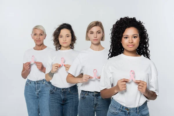 Mulheres multiétnicas em camisetas brancas segurando fitas rosa de consciência de câncer de mama isolado em cinza — Fotografia de Stock