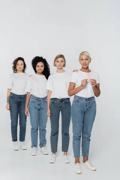 Mujer mayor sosteniendo cinta de la conciencia del cáncer de mama cerca de amigos multiétnicos en el fondo gris - foto de stock