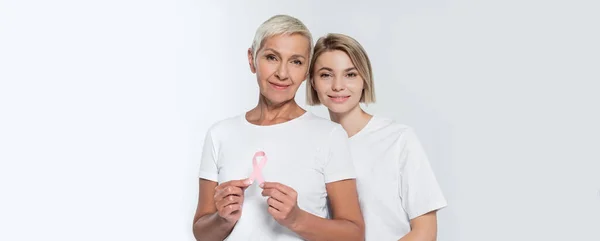 Mujer joven de pie cerca de amigo mayor con cinta de la conciencia del cáncer de mama aislado en gris, pancarta - foto de stock