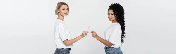 Femmes multiethniques en t-shirts blancs tenant un ruban de sensibilisation au cancer du sein isolé sur une bannière grise — Photo de stock