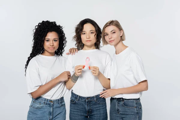 Interracial women hugging friend with ribbon of breast cancer awareness isolated on grey — Stock Photo