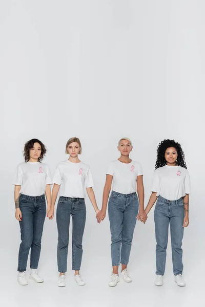 Multiethnic women with ribbons of breast cancer awareness holding hands on grey background — Stock Photo