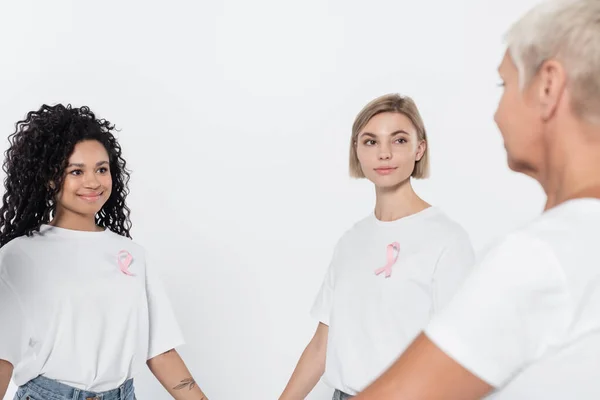 Sorrindo mulheres inter-raciais com fitas de consciência do câncer de mama olhando para amigo sênior isolado em cinza — Fotografia de Stock