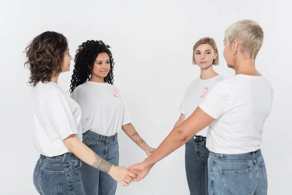 Mujeres multiétnicas con cintas rosas en camisetas tomadas de la mano aisladas en gris - foto de stock