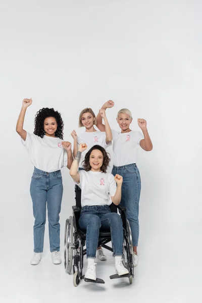 Souriant interracial femmes avec des rubans roses montrant oui geste proche ami en fauteuil roulant sur fond gris — Photo de stock