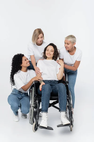 Улыбающиеся многоэтнические женщины, обнимающие друга в инвалидной коляске на сером фоне — стоковое фото