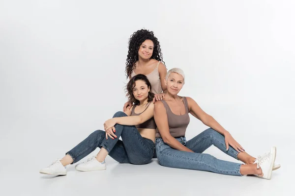 Mulheres positivas sentadas perto de amigo afro-americano em fundo cinza — Fotografia de Stock