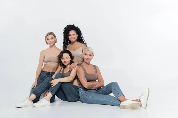 Interracial women smiling at camera while sitting on grey background — Stock Photo