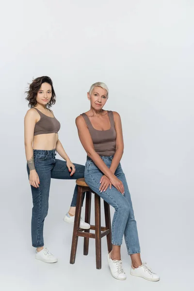 Senior woman sitting on chair near brunette friend on grey background — Stock Photo