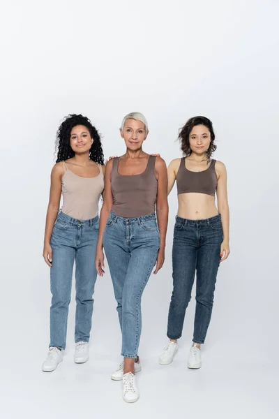 Full length of multiethnic women standing near elderly friend on grey background — Stock Photo