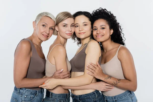 Mujeres multiétnicas abrazando y sonriendo a la cámara aisladas en gris, concepto feminista - foto de stock