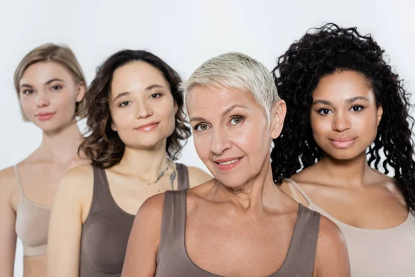 Femme âgée aux cheveux gris souriant à la caméra près d'amis interracial flous isolés sur gris — Photo de stock