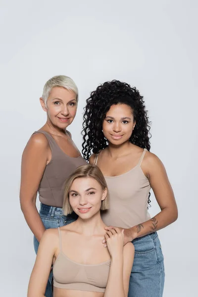 Smiling interracial women looking at camera near young blonde friend isolated on grey — Stock Photo